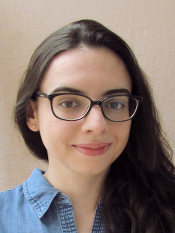 Melissa is a woman from Brazil. She has black long hair, parted to one side. She is wearing a blue denim shirt, a pair of blacck rimmed glasses. She is standing against the wall and smiling while directly looking at the camera.